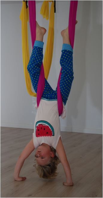 Kinder Yoga Kind hängt kopfüber in bunten Tüchern und trägt ein Shirt mit Wassermelonenmotiv.